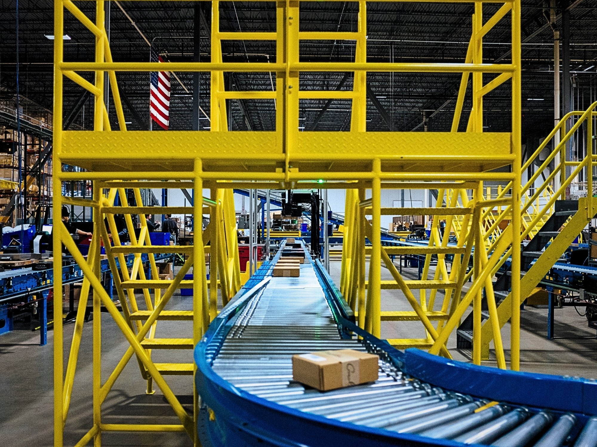 Carton conveyor passing beneath a yellow crossover platform that provides pedestrian access over the conveyor